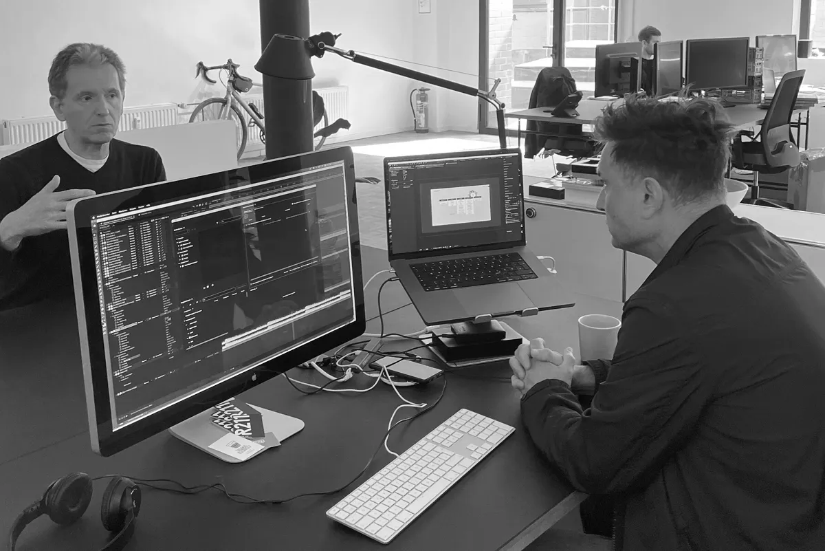 Zwei Personen in einer Marketingagentur sitzen an einem Schreibtisch und besprechen Inhalte vor zwei Monitoren mit geöffneten Projekten und Code-Ansichte