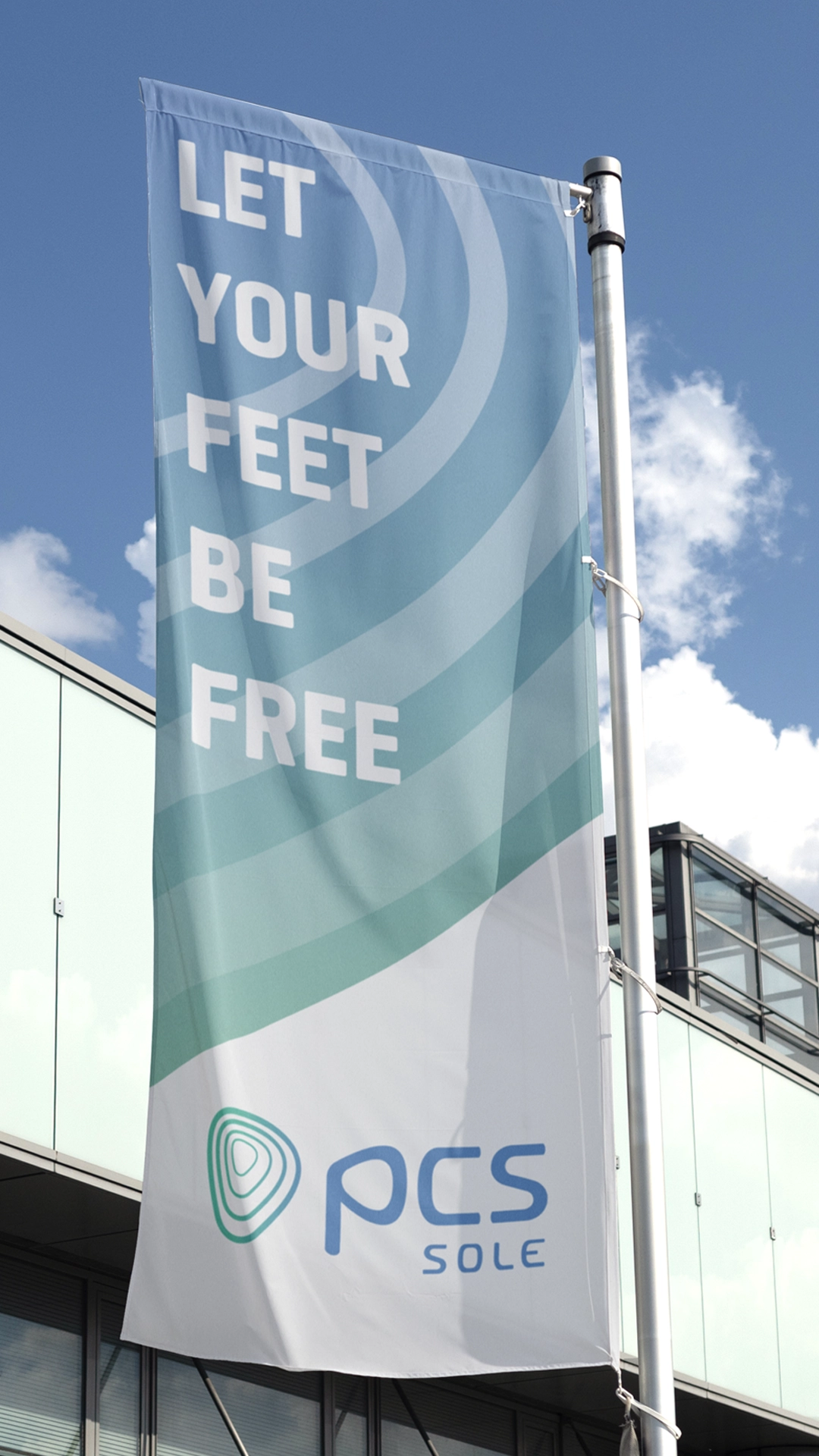 Messefahne zur Vorstellung des Rebandings der Marke PCS-Sole mit dem Claim „Let your feet be free“ und dem Logo von PCS Sole vor blauem Himmel und moderner Glasfassade