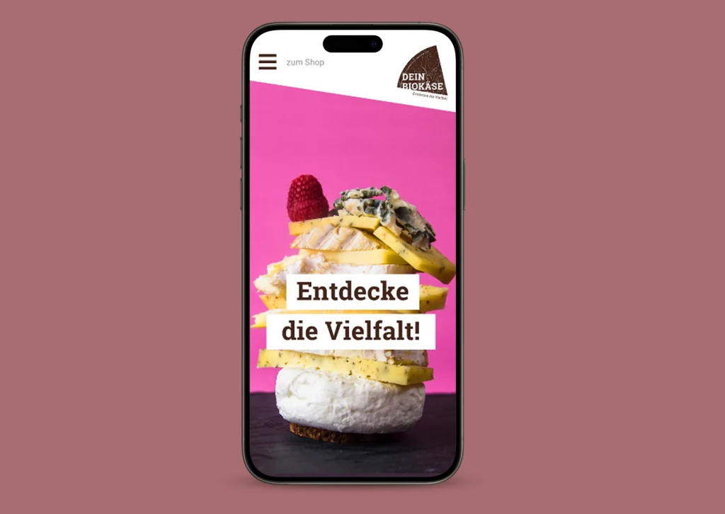 Smartphone mit geöffneter Startseite des DEIN BIOKÄSE Onlineshops, darauf gestapelter Käse mit Himbeere und Claim „Entdecke die Vielfalt“ vor pinkem Hintergrund