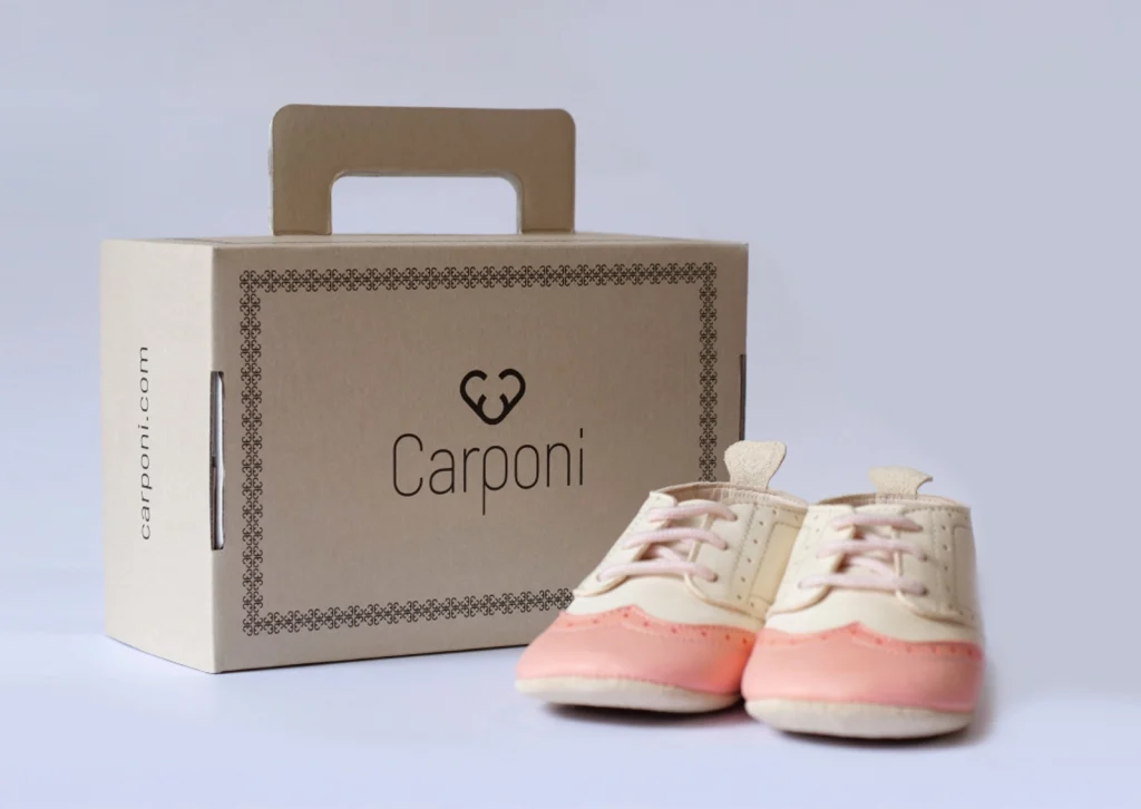 Verpackung und Produkt der Marke Carponi: nachhaltige Krabbelschuhe in Rosé und Beige vor einer Tragebox mit Herzsymbol und Logotyp – Markendesign und Packaging-Gestaltung von R211