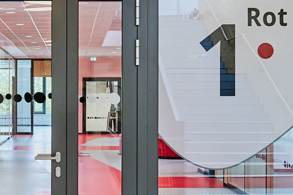 Glaswand mit großflächiger Beschriftung „1. Rot“ im Schulgebäude der Grundschule „Im Pfingsterfeld“ – Teil des farbcodierten Leitsystems von R211 zur Geschoss- und Bereichskennzeichnung im Clusterkonzept