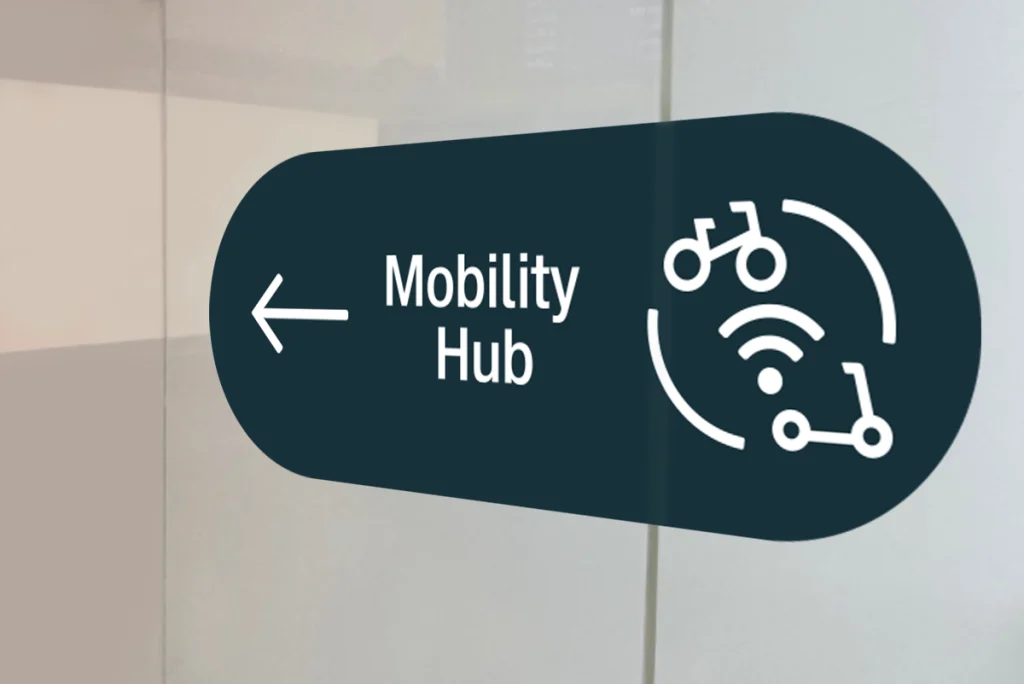 Richtungspfeil mit Mobility-Hub-Beschilderung und Icon für Fahrrad, E-Scooter und digitale Verbindung auf transparenter Tür – Teil des von R211 gestalteten Wegeleitsystems für die Deutsche Bahn
