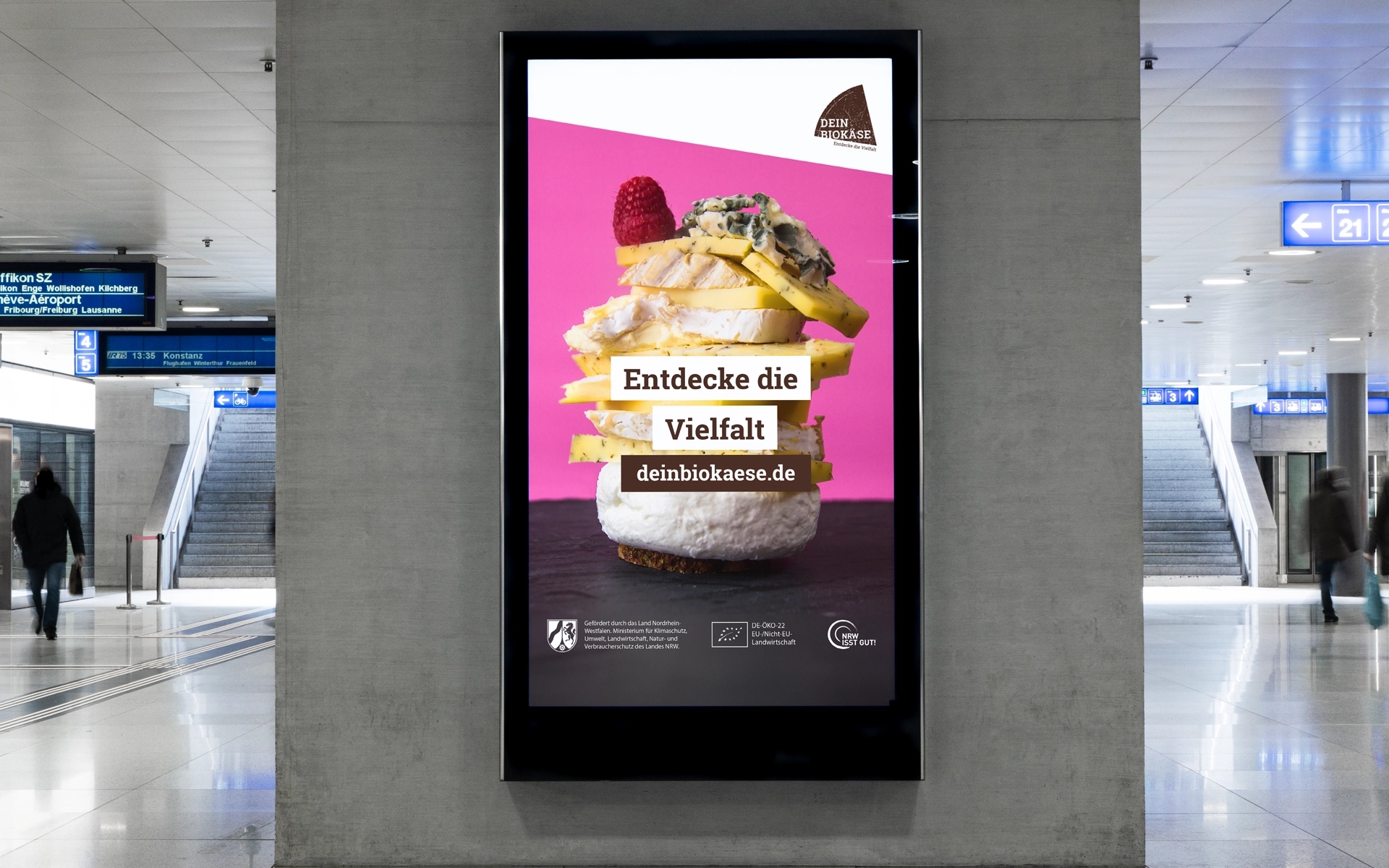 Plakatmotiv von DEIN BIOKÄSE im urbanen Raum, platziert in einem U-Bahnhof: Verschiedene Käsesorten mit Himbeere als Topping auf einer Brotscheibe, inszeniert im modernen Cuisine-Look.