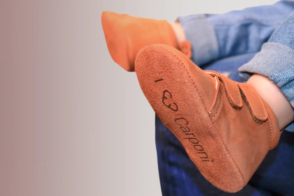 Kinderfuß in cognacfarbenem Carponi-Krabbelschuh mit geprägtem Schriftzug und stilisiertem Herzsymbol auf der Sohle – Markengestaltung und Positionierung durch R211 im Segment nachhaltiger Babyprodukte