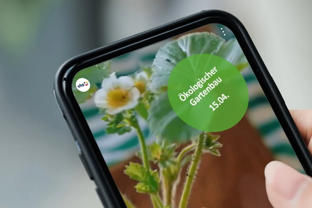 Hand mit Smartphone zeigt Story der vhs-Musterstadt zu ökologischem Gartenbau, grüner Sticker im Rahmen des Social Media Guides des Deutscher Volkshochschulverbands